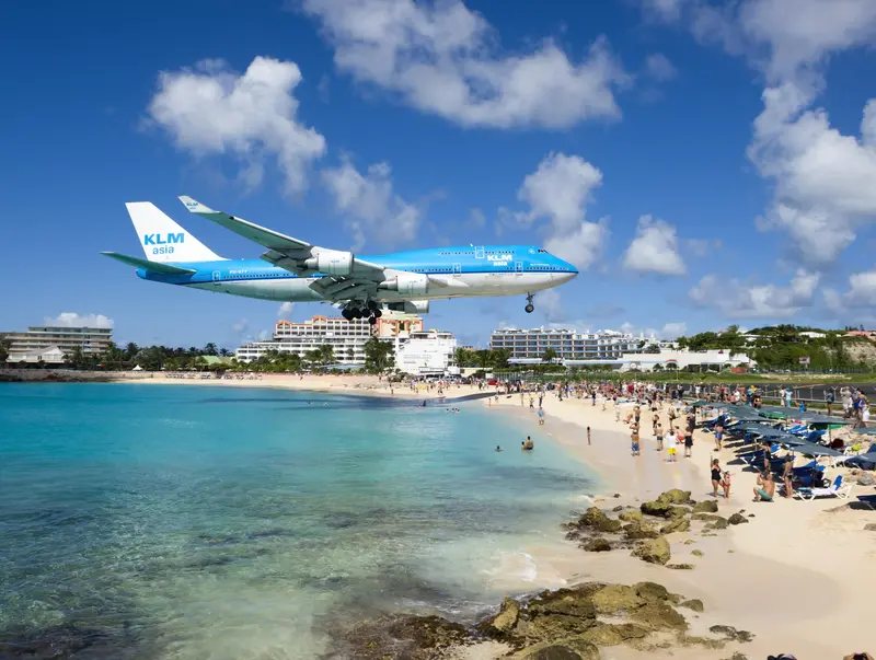 A blue KLM airplane flying low over a crowded beach with clear turquoise waters.