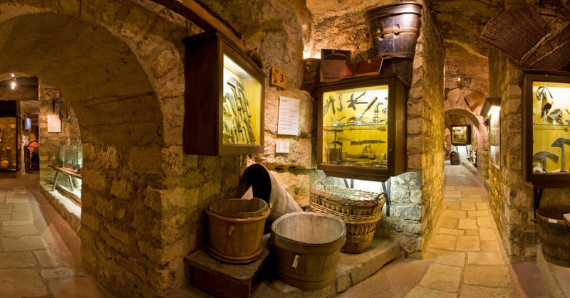 A narrow corridor in the Musée du Vin Paris with stone walls and arches, displaying antique winemaking tools in wooden cases, alongside vintage barrels and woven baskets, evoking the rich winemaking heritage of France.