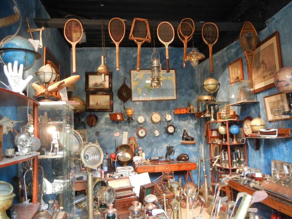 An eclectic collection of vintage objects on display at a Paris flea market, including old tennis rackets, globes, maps, clocks, and scientific instruments, all against a striking blue wall, capturing the allure of unique finds in the city.