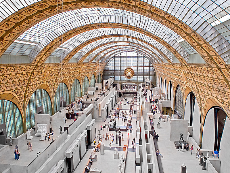 Interior view of the Musée d'Orsay in Paris, showcasing the expansive gallery space with visitors admiring the artworks, beneath the arched, glass ceiling and ornate gold details.