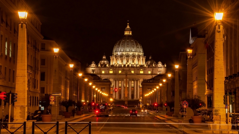 An enchanting Roman night: The Vatican's St. Peter's Basilica, illuminated against the dark sky, marks the end of a street awash in the warm glow of street lamps—inviting explorers to discover the nocturnal magic of Rome.