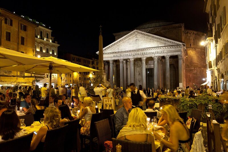 Busy outdoor dining area at night with people seated at tables covered with white cloths, illuminated by soft lights, with the ancient Pantheon in the background, its facade lit and columns distinctly visible, showcasing Rome's vibrant nightlife juxtaposed with historic architecture.