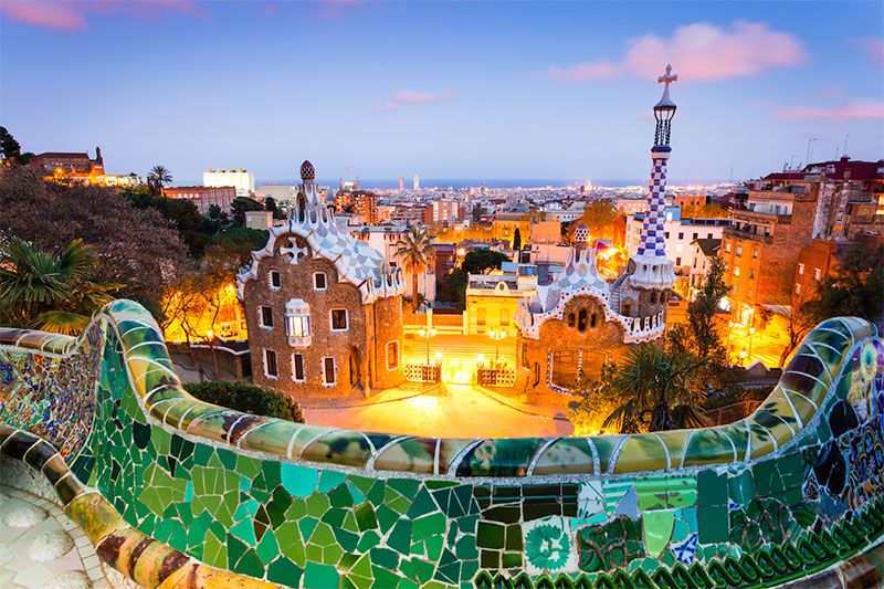 Overlooking the vibrant and whimsical structures of Park Güell in Barcelona, with its unique, trencadís-covered buildings, the serpentine bench, and the iconic mosaic dragon. Visitors stroll along the paths, taking in the panoramic views of the city and the Mediterranean Sea in the distance.
