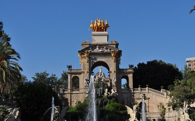 The Cascada Monumental in Ciutadella Park, Barcelona, with its intricate architecture and a golden sculpture of a chariot drawn by horses at the top, set against a clear blue sky, surrounded by lush greenery and water fountains in the foreground.