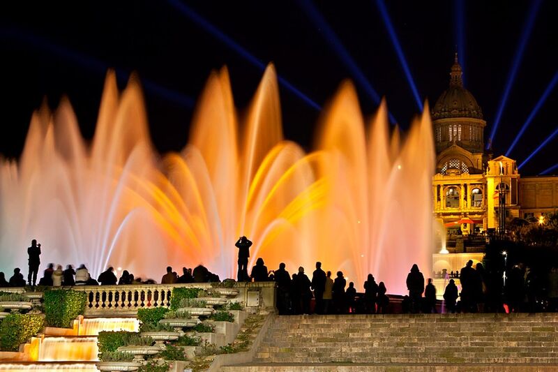 An enchanting night view of the Magic Fountain of Montjuïc in Barcelona, with illuminated, colorful water jets against the backdrop of the National Art Museum of Catalonia, as visitors silhouette along the edges, enjoying the display.