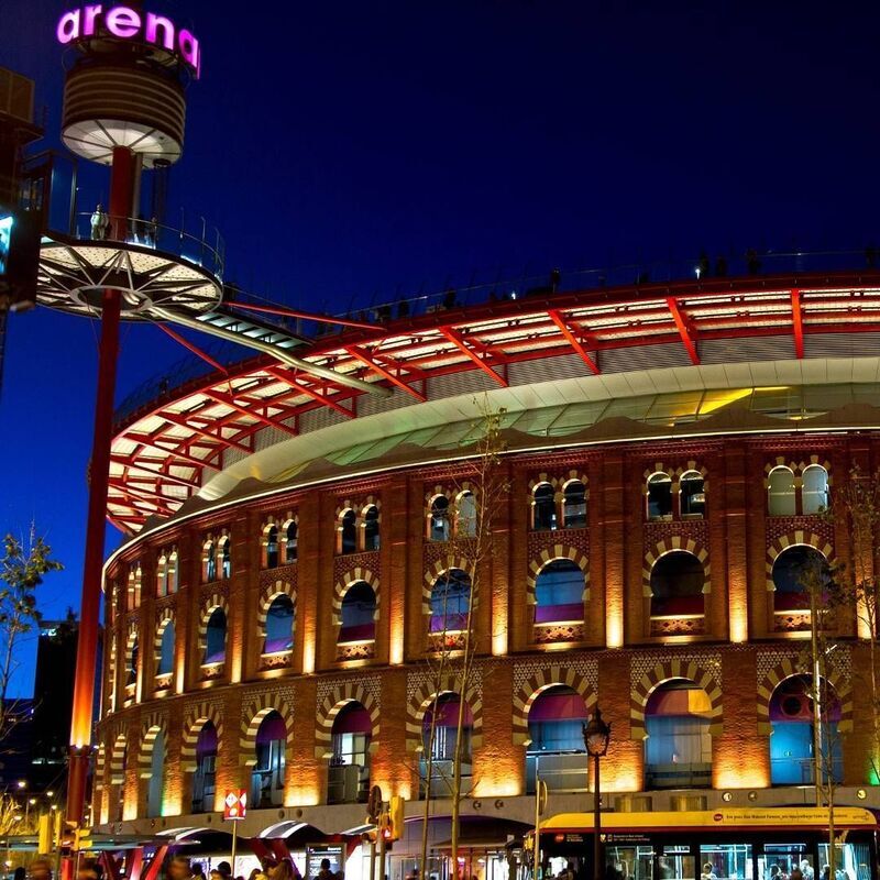Night view of Las Arenas de Barcelona, a former bullring converted into a contemporary shopping mall, with its traditional red brick facade illuminated and topped by a sleek circular structure, blending the city's historic past with its vibrant present.