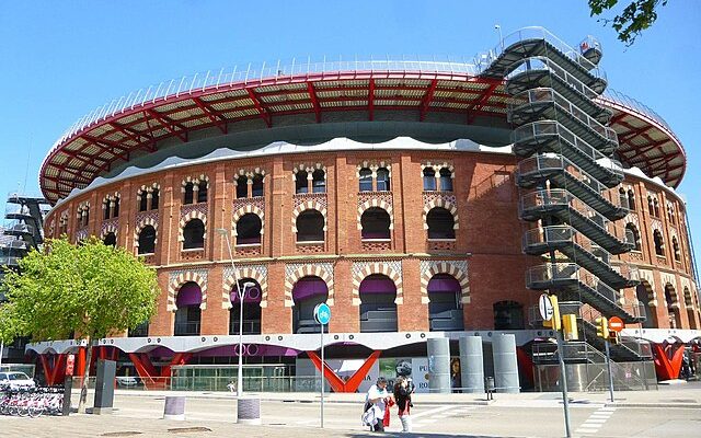 The distinctive red brick facade of Las Arenas, a former bullring turned shopping center in Barcelona, featuring a circular design with a contemporary rooftop structure. Pedestrians are seen walking by, illustrating the blend of historic architecture and modern urban life.