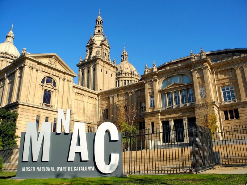 The grand facade of the National Art Museum of Catalonia, with its large 'MNAC' sign in the foreground, stands under a clear blue sky. The museum's Renaissance architecture, featuring domes and detailed stonework, showcases the cultural heritage of Barcelona.