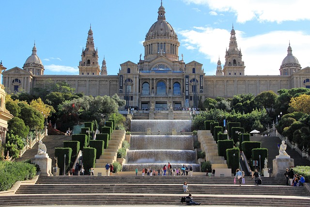 The grand National Art Museum of Catalonia, poised atop Montjuïc Hill in Barcelona, overlooks cascading waterfalls and staircases. Visitors are scattered on the steps, enjoying the view under a bright blue sky, with lush greenery accentuating the scene.