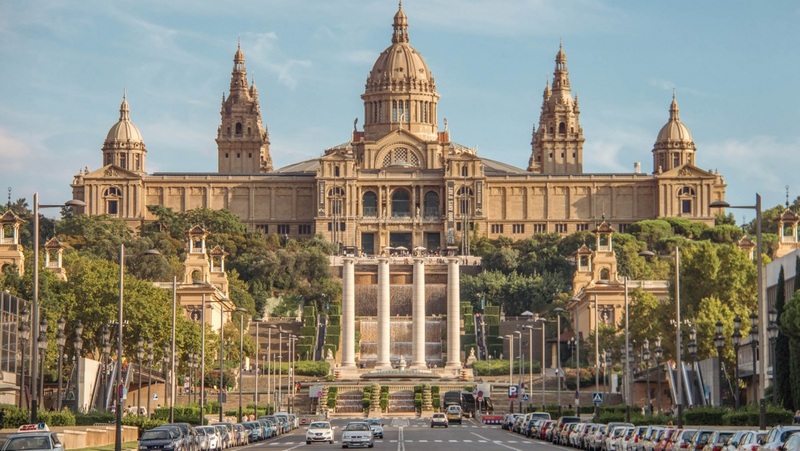 Photograph of the Palau Nacional in Barcelona, showcasing its impressive architecture with two towering spires and a large central dome, set against a blue sky. The foreground features a broad avenue leading up to the museum, lined with parked cars and lush greenery on the sides.