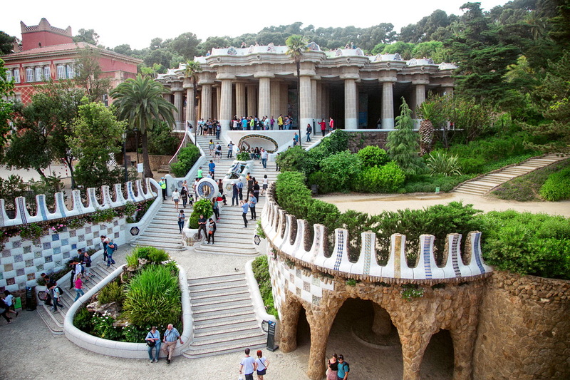 Tourists meandering through the iconic serpentine bench and the columned hall of Park Güell, one of Antoni Gaudí's most famous creations in Barcelona, with rich vegetation and unique architectural elements under a clear sky.