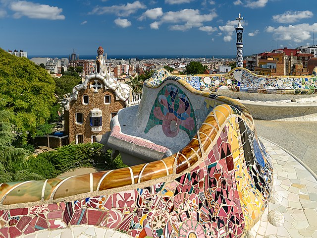A sweeping view from Park Güell's main terrace, with its iconic mosaic serpentine bench in the foreground, curving around the edge. The vibrant mosaics glisten in the sunlight, with Gaudí's whimsical architecture and the cityscape of Barcelona stretching out towards the sea in the background.