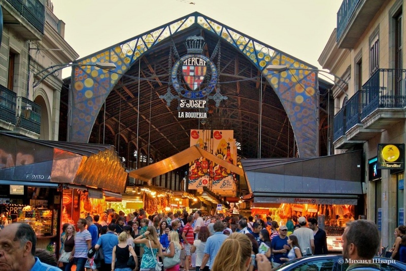 Crowds of locals and tourists alike flock to the entrance of Mercat de Sant Josep de la Boqueria in Barcelona, a bustling market renowned for its fresh produce and vibrant atmosphere, with the market's colorful sign and mosaic decorations under a dusk sky.