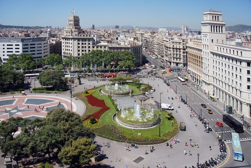 Bird's eye view of Plaça de Catalunya, a large and vibrant public square in Barcelona. The scene is busy with people and traffic, the movement captured around the central fountains and landscaped gardens, framed by grand buildings under a clear blue sky.