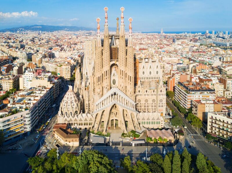 Aerial view of La Sagrada Família, the masterpiece of architect Antoni Gaudí, in Barcelona, with its intricate spires towering over the cityscape, showcasing the blend of Gothic and Art Nouveau styles against the canvas of the urban grid.