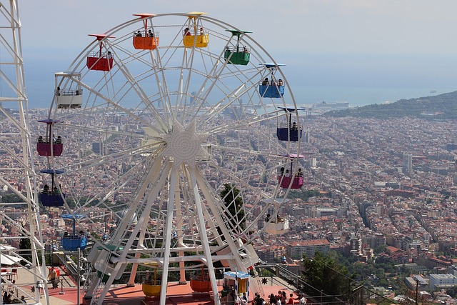 Colorful cabins of a Ferris wheel stand out against the panoramic backdrop of Barcelona's urban landscape, viewed from the summit of Tibidabo Mountain. The sprawling city and the Mediterranean Sea in the distance create a stunning contrast with the amusement ride's playful presence.