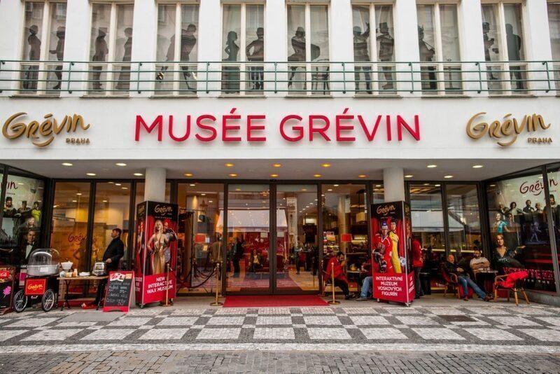 The entrance to Musée Grévin in Paris, inviting visitors inside with its bold red signage above the door and a plush red carpet leading the way. Life-size wax figures can be glimpsed through the glass façade, teasing the lifelike encounters that await within this renowned wax museum.
