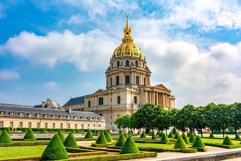 The majestic Hôtel National des Invalides under a bright blue sky with fluffy clouds, featuring its iconic golden dome shining brilliantly. In the foreground, geometrically shaped hedges in lush gardens provide a serene and ordered contrast to the grandeur of the historic building.