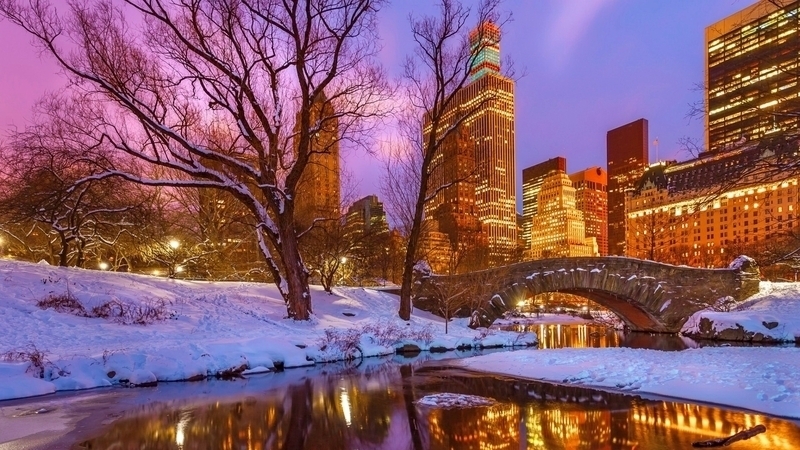 A serene winter scene in Central Park at twilight with snow-covered grounds and a stone bridge, reflected in the still waters, with the city's illuminated skyline providing a stunning backdrop for Christmas activities in New York.





