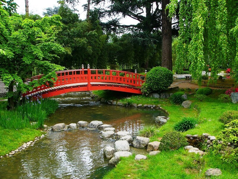 Vibrant red Japanese-style bridge arching over a tranquil stream in the lush greenery of the Albert Kahn Museum and Gardens, with weeping willows and flowering shrubs adding to the serene landscape.
