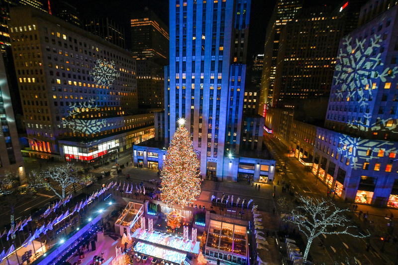 A breathtaking nighttime overview of Rockefeller Center during Christmas, with the radiant Christmas tree at its heart and snowflake projections on surrounding buildings, showcasing a top holiday activity in New York.