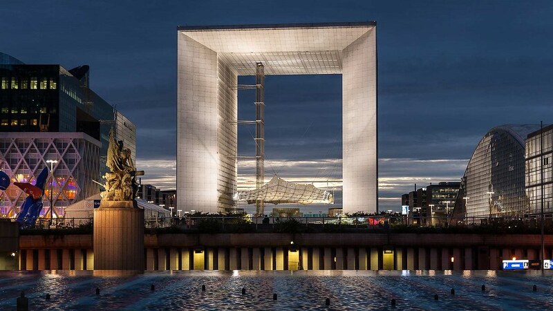 Twilight view of the Grande Arche in La Défense, Paris, with its imposing modern architecture and a statue in the foreground, reflecting in the water below and surrounded by illuminated office buildings.