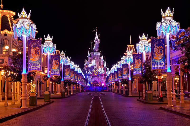 Main Street in Disneyland Paris illuminated at night with vibrant lights and festive decorations, leading towards the iconic Sleeping Beauty Castle adorned with twinkling lights and a starry sky above.