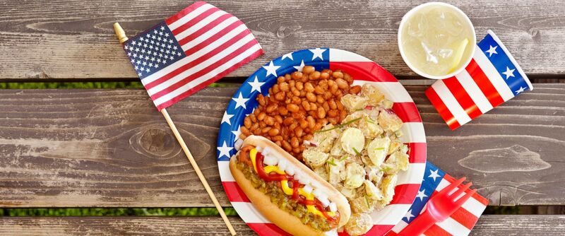 A traditional American meal featuring a hot dog, baked beans, potato salad, and a lemonade, served on a patriotic-themed plate with American flags, capturing the culinary delights to enjoy while visiting New York City.