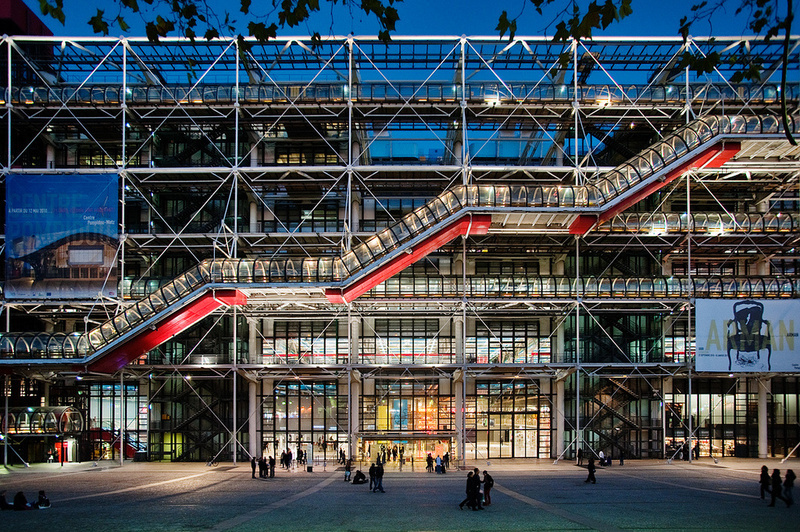 The Centre Pompidou in Paris captured at dusk, showcasing its iconic exterior escalators and structural skeleton, with visitors milling around the entrance.