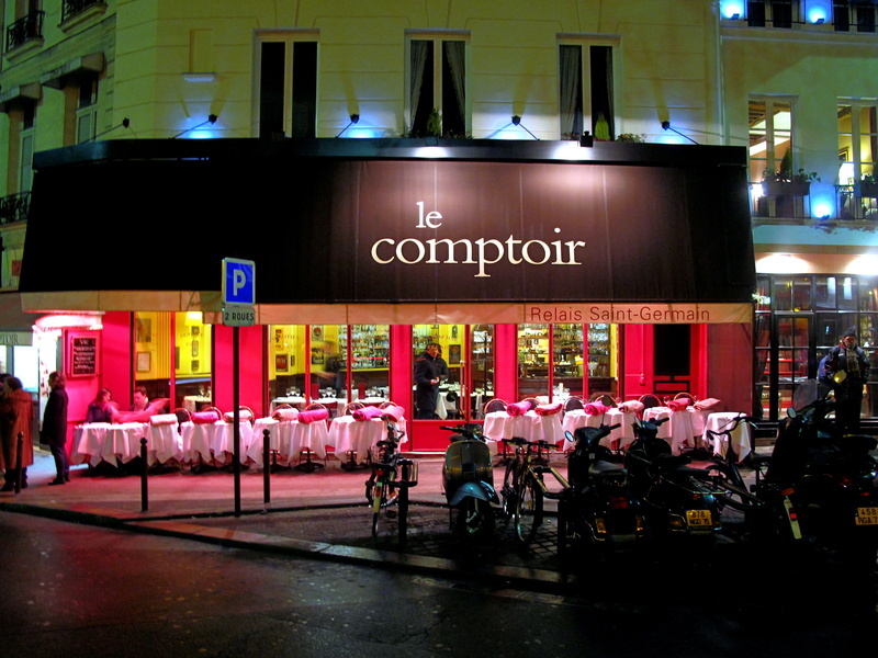 The vibrant and inviting exterior of Le Comptoir du Relais in Paris at night, with its iconic red awning and neatly arranged outdoor seating, bustling with patrons and surrounded by the warm glow of streetlights, capturing the essence of the city's cherished bistro culture.