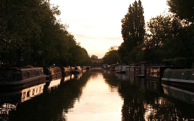 A serene sunset over Little Venice in London, with the glow of the evening sky reflecting on the calm canal waters, lined by moored narrowboats and bordered by lush trees.