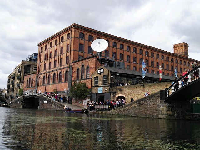 A bustling scene at Camden Market by the canal, with people gathered at outdoor tables under the shadow of an imposing brick building.