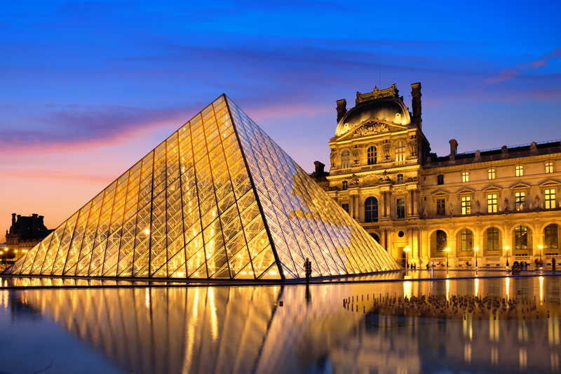 The Louvre Pyramid glows like a jewel against the dusk sky, with its glass panes reflecting the sunset's warm hues. The historic Louvre Palace looms gracefully in the background, its classical architecture illuminated by golden lights, while the pyramid's reflection ripples softly in the courtyard's still water.