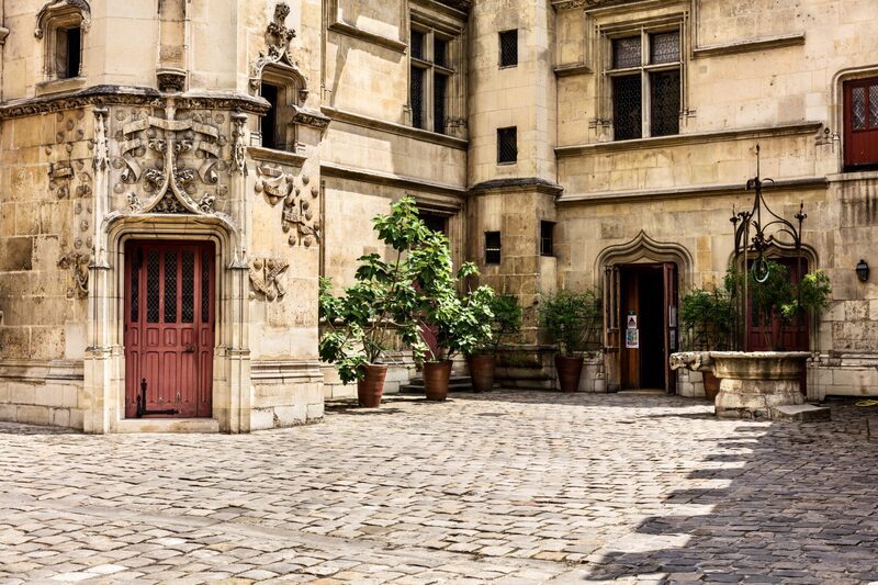 A secluded cobblestone courtyard tucked away in the Marais district of Paris, flanked by historical buildings with ornate stonework and wood-framed windows. Potted green plants add life to this serene, hidden nook, inviting passersby to ponder the tales it could tell.