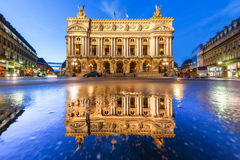 Twilight descends on the Opéra Garnier in Paris, its elaborate façade illuminated against the deepening blue sky. The building's reflection is mirrored perfectly on the wet street surface below, adding to the scene's symmetrical beauty. City lights and the early evening bustle give a lively contrast to the calm elegance of the opera house.