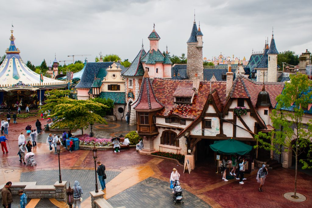 View of Fantasyland in Disneyland Paris, featuring storybook architecture with colorful facades and pointed roofs, visitors wandering the cobblestone streets, and a carousel in the background under a cloudy sky.