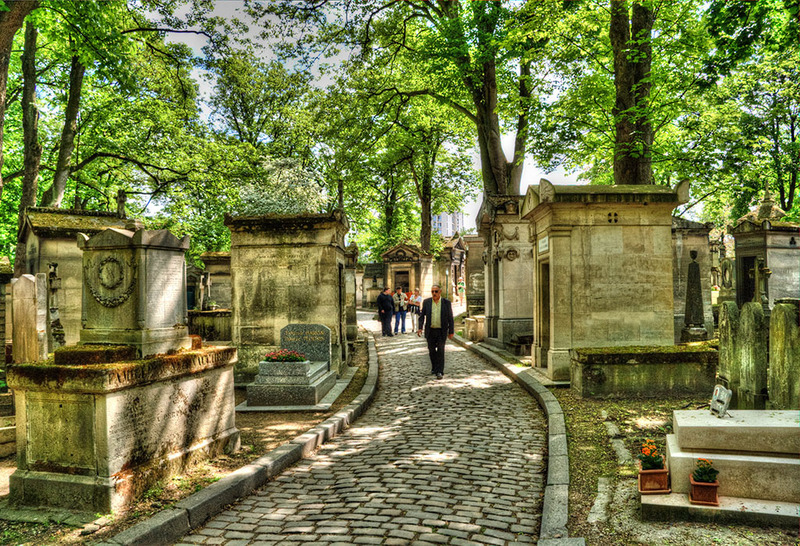 Visitors walk along a cobblestone path in Pere Lachaise Cemetery in Paris, surrounded by a variety of ornate tombstones and lush green trees, with a clear sky above.