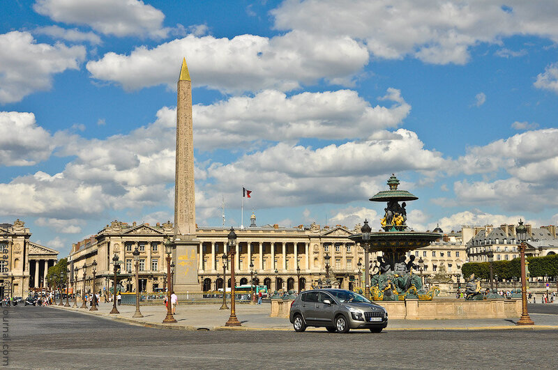 The Place de la Concorde on a sunny day, characterized by the ancient Egyptian obelisk standing tall at its center, flanked by two ornate fountains. The grand buildings that line the square add a touch of Parisian elegance.