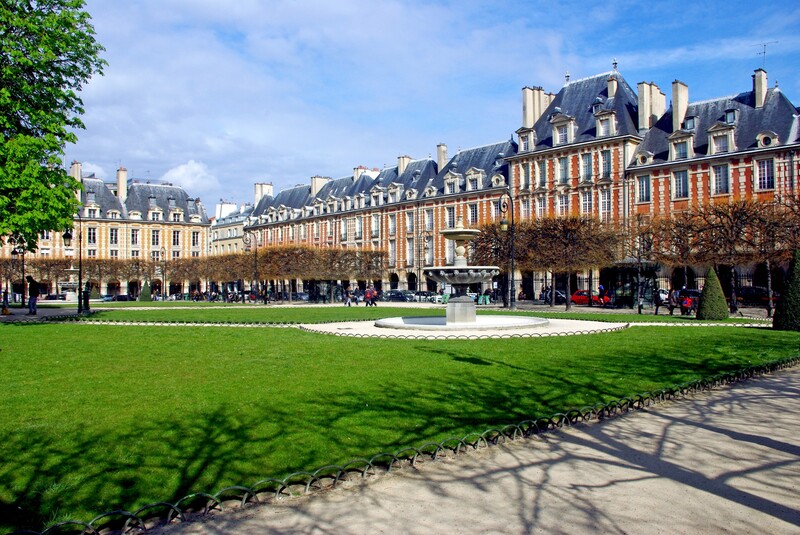 The historic Place des Vosges on a sunny day, with lush green lawns, a central fountain, and lined with symmetrical red brick facades and slate roofs, as people enjoy a leisurely day in one of Paris' most charming squares.