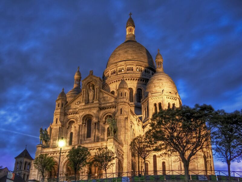 The Sacré-Cœur Basilica, perched atop Montmartre, is aglow with warm lighting against the backdrop of a dramatic twilight sky. The basilica's white domes and intricate architecture are highlighted, with silhouettes of trees framing the scene, capturing the serene yet majestic ambiance of this iconic Parisian landmark.