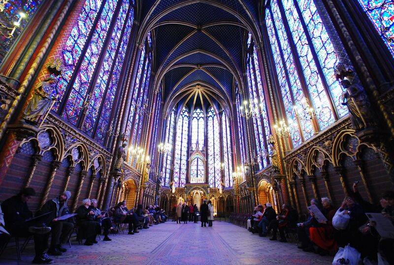 The interior of Sainte-Chapelle in Paris, showcasing its towering Gothic architecture with a vaulted ceiling and majestic stained glass windows in rich blues and reds. Visitors sit along the sides, immersed in the chapel's tranquil beauty, with elegant chandeliers illuminating the space.
