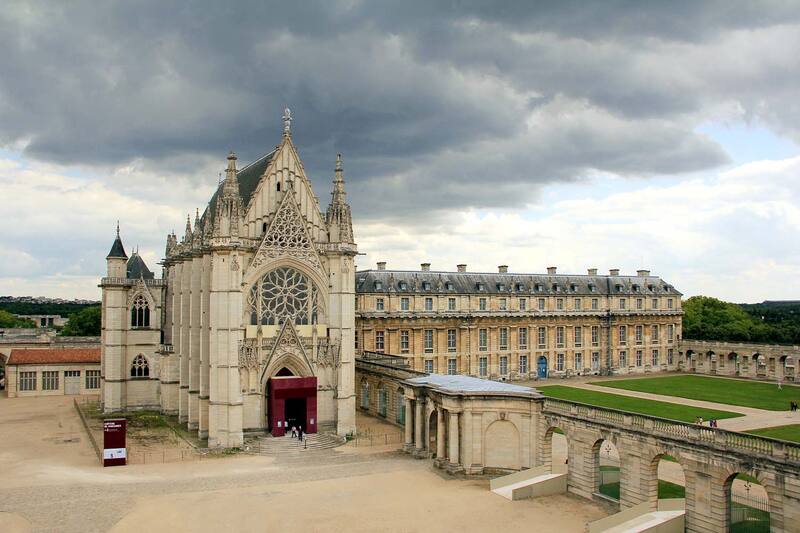The striking Chapel of Château de Vincennes under a dramatic cloudy sky, showcasing intricate Gothic architecture with a grand entrance and delicate spires. The chapel stands prominently in the foreground with the expansive château stretching out behind it, encircled by an open courtyard.