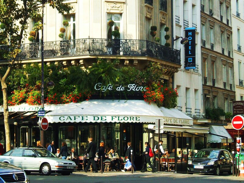 The bustling exterior of Café de Flore in Paris, adorned with vibrant red flowers, with patrons seated outside. The historic café's façade is complemented by a traditional Parisian building with ornate balconies above, and a blue 'HOTEL' sign visible next door. Pedestrians and cars pass by, capturing a lively Parisian street scene.