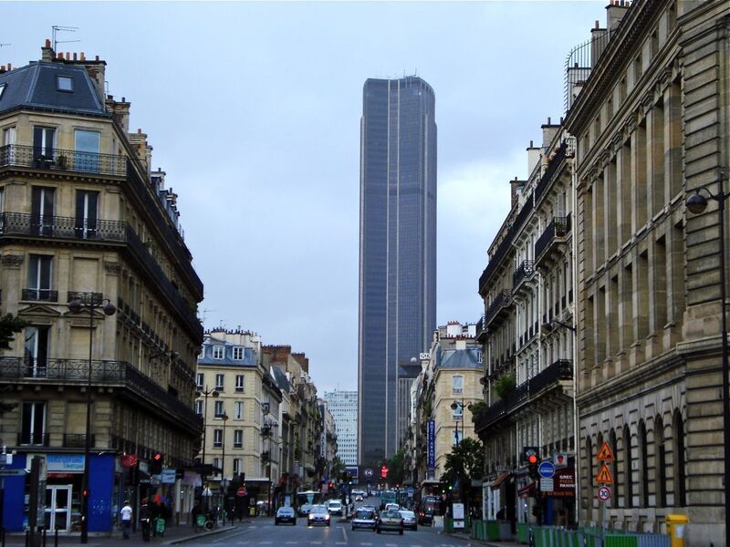 The view along a typical Parisian street leading to the towering Montparnasse Tower under an overcast sky, with traditional buildings lining the street and daily city life unfolding below.