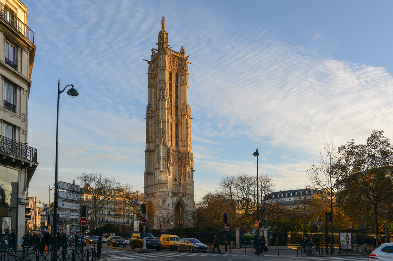 Image of the historic Saint-Jacques Tower standing tall amidst the hustle of Parisian streets, with pedestrians and traffic under a sky streaked with clouds, encapsulating the blend of history and modernity for travelers.