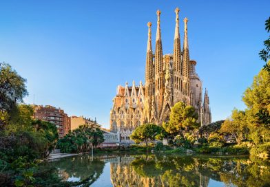 The La Sagrada Família basilica in Barcelona, captured on a clear day, with its intricate Gothic and Art Nouveau façades and towering spires reflecting in the pond in front, surrounded by lush greenery.