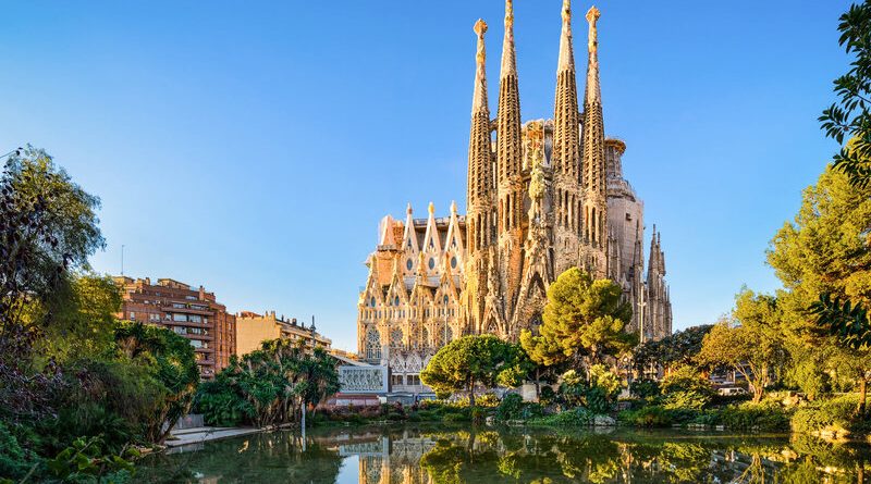 The La Sagrada Família basilica in Barcelona, captured on a clear day, with its intricate Gothic and Art Nouveau façades and towering spires reflecting in the pond in front, surrounded by lush greenery.