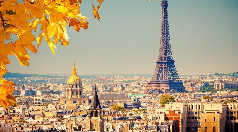 A panoramic view of Paris in autumn, showcasing the Eiffel Tower and the golden dome of Les Invalides amidst a sea of traditional Parisian buildings. The foreground features vibrant yellow autumn leaves, suggesting the photo was taken from a high vantage point through the foliage, on a clear day with a bright blue sky.
